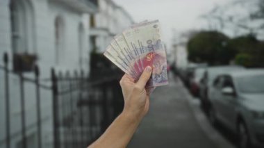 Şehir sokaklarında Arjantin pesoları tutan adam, canlı banknotları bulanık bir şehir geçmişine karşı sergiliyor, para birimini ve ekonomik temaları vurguluyor..