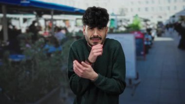 Restoran terasında elleri soğuk sokakta birbirine yapışmış genç bir adam süveter giymiş rahatsız görünüyor..