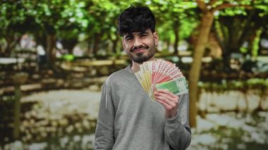 Elinde Norveç kronu banknotları tutan bir adam açık hava parkında gülümsüyor yemyeşil ve doğal aydınlatma ile huzurlu çevreyi vurguluyor..