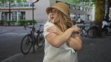 Sarışın, omuzlarını kenetlenmiş bisiklet ve park etmiş scooterların yakınındaki bir caddede kendine sarılan bir kadın, endişe dayanıklılığı..