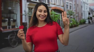 Sol elinde küçük kırmızı bir kalp tutan ve bir şehir caddesinde üç parmağını gösteren bir kadın gülümsüyor..