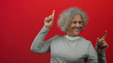 Canlı kırmızı bir duvara karşı neşeyle dans eden kadın, enerjik duruşu ve etkileyici gri saçlı görüntüsüyle mutluluğunu ve güvenini sergiliyor..