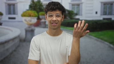 Gözlüklü genç İspanyol adam avucunu kaldırıyor ve şüpheli bir şekilde sokaktaki kameraya bakıyor..