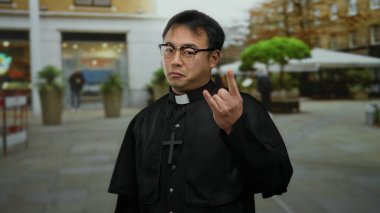 Gözlüklü bir adam ve haç kolyeli bir rahip binaların ve yeşilliklerin bulanık arka planına sahip bir şehir sokağında duruyor..