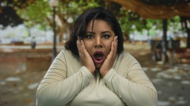 Açık hava parkında, ağaç gölgesinin altında şaşkınlık ve duygu gösteren, şok olmuş yüz ifadesi olan genç ve iri bir kadın.