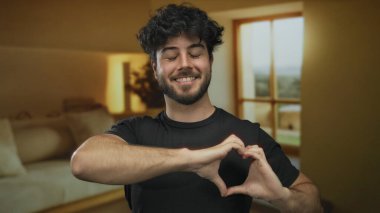 Elleri rahat otel odasında, geniş pencereli ve sıcak ışıklandırmalı kalp sembolü yapan genç adam mutluluğu ve olumlu duyguyu ifade ediyor..