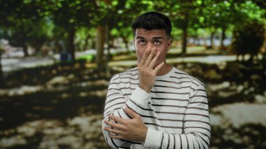 Çizgili tişörtlü İspanyol adam gölgeli ağaçlarla kaplı güneşli bir parkta şaşırmış görünüyor..