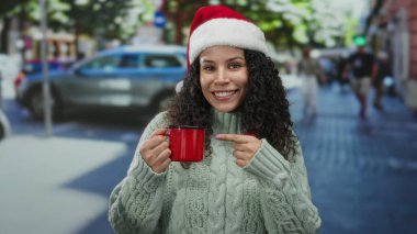 Noel Baba şapkalı İspanyol kadın kırmızı bardağı işaret ediyor. Kalabalık bir caddede, rahat bir kazak giyiyor. Şenlikli şehir tatil temasını vurguluyor..