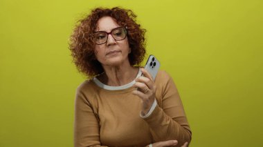 Orta yaşlı esmer kadın, kolları çaprazlanmış, izole edilmiş sarı arka plana karşı akıllı telefon tutuyor..