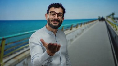 Sakallı İspanyol bir adam, neşeli bir sahil yürüyüşü sahnesini somutlaştıran canlı bir mavi denize ve berrak gökyüzüne karşı bir öpücük yolluyor..