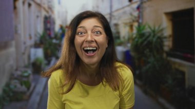 Orta yaşlı, iri gözlü, eli göğsünde ve ağzı açık bir kadın dar sokakta şaşkınlığını ifade ediyor; neşe.