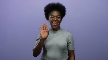 Mor arka planda selamlaşan kadın, şakacı bir şekilde dilini uzatıyor ve göz kırpıyor; genç, neşeli, Afrikalı, Amerikalı kadın sıcaklık ve dostluk saçıyor..