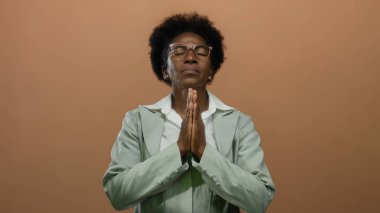 Elleri sımsıkı bağlı, gözlüklü bir kadın, tenha kahverengi bir arka plana karşı duruyor, huzurlu bir duygu ve inanç iletiyor, huzurlu, yansıtıcı bir ortama.