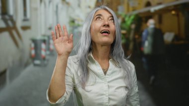Gri saçlı, gülümseyen ve canlı bir şehir sokağında el sallayan bir kadın şehir ortamında kendine güvenini ve sıcaklığını gösteriyor..