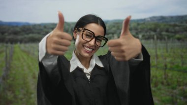 Yargıç üniforması içinde gülümseyen bir kadın, manzaralı bir parkta başparmak kaldırıyor, mutluluk ve profesyonelliği canlı yeşil bir arka planda somutlaştırıyor..