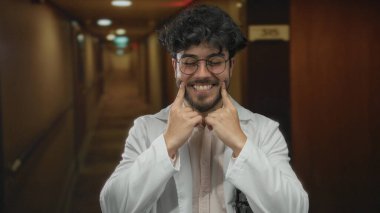 Otel koridorunda gülümseyen, sakallı ve gözlüklü, mutluluğu ve İspanyol kültürünü yansıtan kapalı mekan modasını tasvir eden genç bir adam..