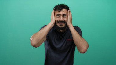 Sıkıntılı sakallı İspanyol bir adam canlı yeşil arka planda başını tutuyor, hayal kırıklığını ve kafa karışıklığını ifade ediyor..