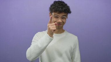 Gözlüklü ve beyaz kazaklı genç İspanyol adam mor stüdyoda kamerayı işaret ediyor..