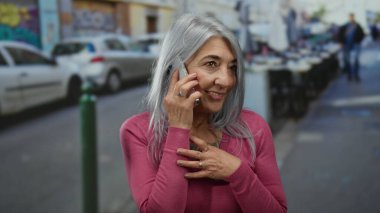 Kentsel sokak ortamında gülümseyen bir gülümseme ile konuşan bir kadın gri saçlı ve pembe kazaklı yaşlı bir kadını günlük ortamda açık havada gösteriyor..