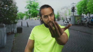 Neon renkli sarı tişörtlü adam şehir caddesindeki kamerayı işaret ediyor. Avuç içi ve kol dışa doğru uzanıyor..