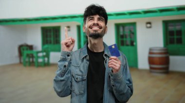 Yeşil dekorlu, açık hava restoranının terasında kredi kartıyla gülümseyen bir adam güneşli günün tadını çıkarıyor..