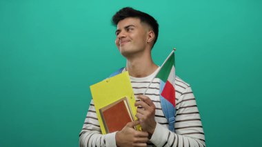 Elinde İtalyan bayrağı ve çizgili gömlek ve sırt çantası olan bir adam izole edilmiş yeşil arka plana karşı düşünceli bir ifadeyle ayakta duruyor..