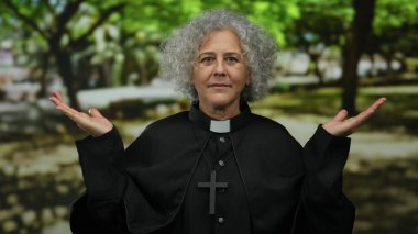 Gri saçlı, papaz kıyafeti giymiş yaşlı bir kadın sakin bir açık hava parkı ortamında duruyor. Dalgınlığı ve maneviyatı yemyeşil ağaçların altında somutlaştırıyor..