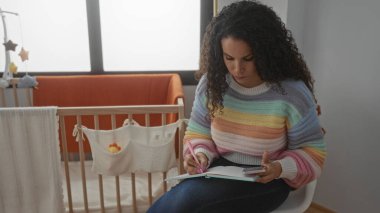 Siyah kıvırcık saçlı genç bir kadın, huzurlu bir ev sahnesini beşikteki bebek karyolasının yanına oturmuş, bir deftere yazıyor..