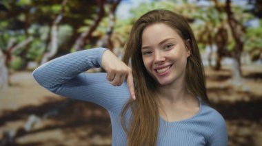 Mavi tişörtlü genç kadın gülümsüyor ve parmakla işaret ediyor güneş ışığı altındaki yeşil orman ağaçlarının arasında; mutluluk.