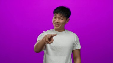 Genç Çinli bir adam, beyaz bir gömlek giyerken izole edilmiş pembe bir arka plan üzerinde neşeli jestler yapıyor, samimi ve esprili bir hava yayıyor..