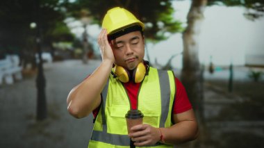 Şehir caddesinde kahve tutan inşaat işçisi. Kulaklık ve güvenlik yeleği takıyor. Yorgun bir ifadeyle..