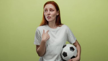 Sarı bir duvara karşı şaşkın bir ifadeyle futbol topu tutan bir kadın, izole bir arkaplanda kırmızı saç ve günlük beyaz gömlek.