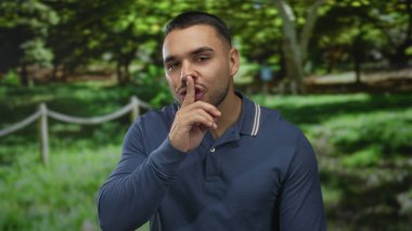 Yeşil ormanda sessizlik için parmaklar dudaklara mavi polo gömlek giyiyor ve güneş ışığı altında açık havada gülümsüyor; iç sessizlik.