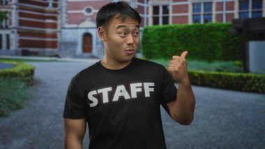 Personel gömlekli genç adam, kırmızı tuğla bina duvarı ve manikürlü çitlerin önünde baş parmağını kaldırıyor. Onaylandı..