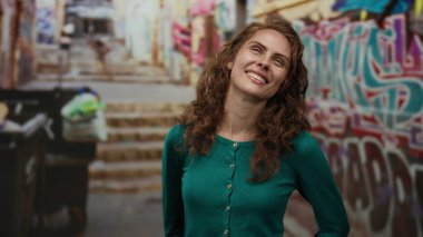 Grafiti dolu şehir sokaklarında gülümseyen bir kadın, yeşil bir kazak giymiş, gözleri kapalı neşeli ve tasasız bir yüz ifadesi sergiliyor..