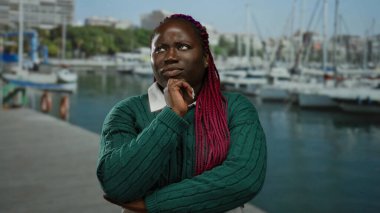 Limanda arka planda tekneler ve denizle kafa yoran bir kadın, açık deniz ortamında düşünceli bir yüz ifadesi sergiliyor..