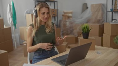 Yeni evde dizüstü bilgisayar kullanan bir kadın. Oturma odasındaki kutularla çevrili. Kulaklık takmış, günlük kıyafetler giyiyor..