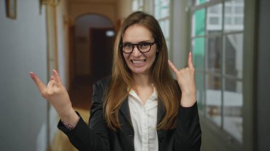 Genç bir kadın dilini uzatır ve parlak bir otel koridorunda kaya boynuzları hareketi yapar. Enerji heyecanı..