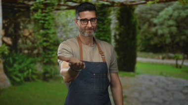 Adam, kot önlüklü ve sakallı gözlüklü olarak parkta elini uzatır; cömertliği ve nezaketi hoş karşılar..