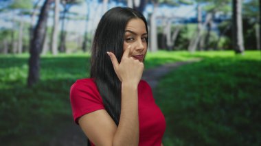 Kırmızı tişörtlü İspanyol kadın, uzun ağaçlarla çevrili yemyeşil bir parkta kendine güvenen bir ifadeyle bir jest yapıyor..