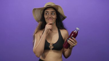 Kadının elinde mor stüdyoda hasır şapka ve siyah bikinili ferahlatıcı meyve suyu şişesi var..