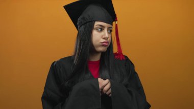 Mezuniyet üniformalı genç İspanyol kadın turuncu arka planda yalnız duruyor ve hayal kırıklığı yaşıyor..