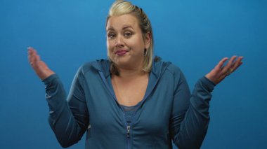 Stüdyoda gözleri kapalı dururken mavi kapüşonlu genç sarışın kadın omuz silkiyor; belirsizlik.