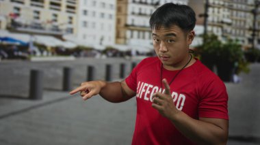 Genç cankurtaran, elinde ciddi bir bakışla ıslık tutarken işlek şehir caddesi binasını işaret ediyor; tetikte olma görevi.