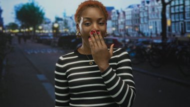 Sessizlik için parmakları dudağında olan kadın alacakaranlıkta şehir caddesinde duruyor çizgili süveter ve altın küpeler takıyor; sessizlik..