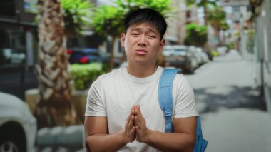 Sırt çantalı genç bir adam güneşli bir şehir caddesinde durup dua ederken palmiye ağaçları ve park halindeki arabaların arasında duygularını ifade ediyor..