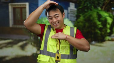 Sarı yelekli bir adam arka planda ağaçlar ve binalar olan şehir sokağında gülümsüyor ve Asyalı yetişkin bir erkeğin yüksek görüş mesafesinde olduğu açık hava sahnesini çekiyor..