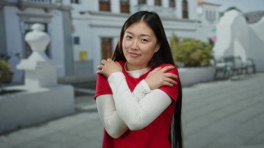 Kırmızı giyinmiş, açık havada gülümseyen, kendini sıcak tutan, güneşli bir sokakta mutluluk ve memnuniyet gösteren bir kadın..