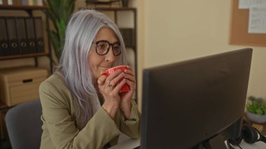 Ofiste çalışan, elinde kırmızı kupayla bilgisayar kullanan, modern bir çalışma alanında profesyonel ve düşünceli bir tavır sergileyen yaşlı bir kadın..