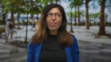 Kahverengi saçlı, mavi ceketli ve gözlüklü bir kadın yeşil ormanda dilini uzatır, kaygısızca yaramazlık yapar..